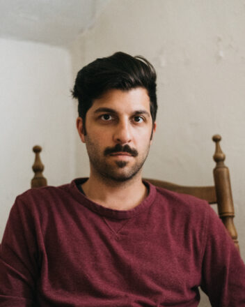 A 40 something white man of Italian descent sitting in a wooden high back chair in front of textured white walls looks intensely into the camera. He's wearing a red long sleeved t-shirt, has short wavy dark hair and a dark moustache with a stubble beard.