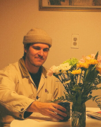 A stylized grainy image of a 30-year-old white man sitting at a kitchen table holding a mug in front of a vase of tall, colourful flowers. Evan wears a light coloured work jacket and toque, has prominent dark eyebrows and is looking down at the flowers while softly smiling.