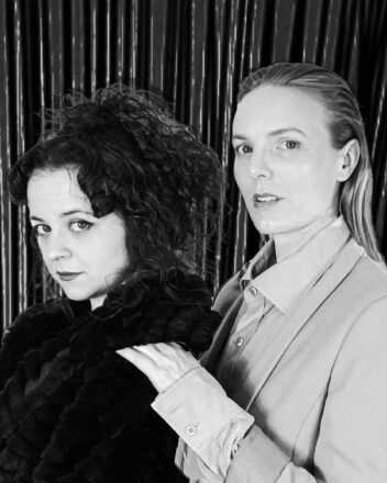 A black and white photo of two white women standing in front of a curtain backdrop. Lauren, who is shorter and has long dark curly hair and wears a lush black fur coat, stands in front of Alaine, who has long straight blonde hair and wears a light coloured collared shirt and suit jacket. Their shoulders face camera left and their heads are turned to look directly into the camera with an invitational stare.